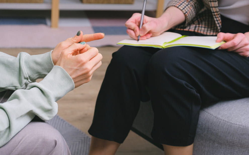 Two people in a therapy session, one gesturing while explaining something and the other taking notes in a notebook